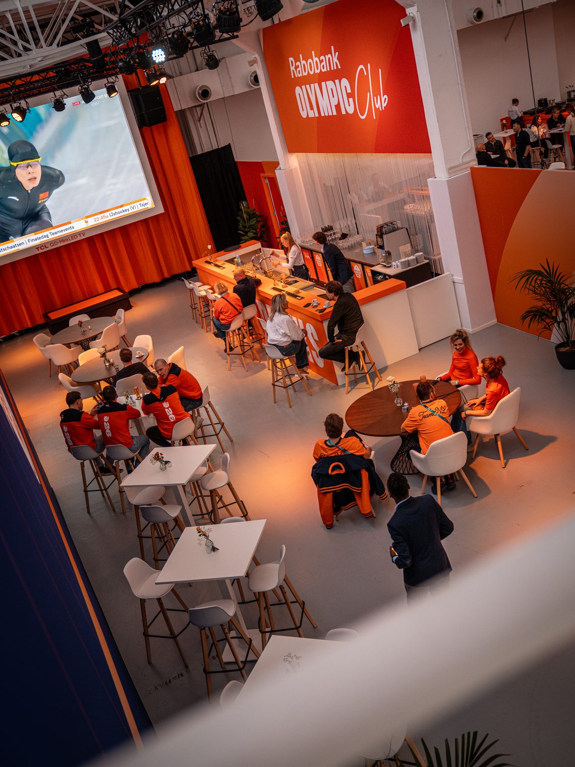 Crew en bezoekers in oranje aan de bar en tafels van het Staatsloterij TeamNL Huis, met koffiemachines achter de bar
