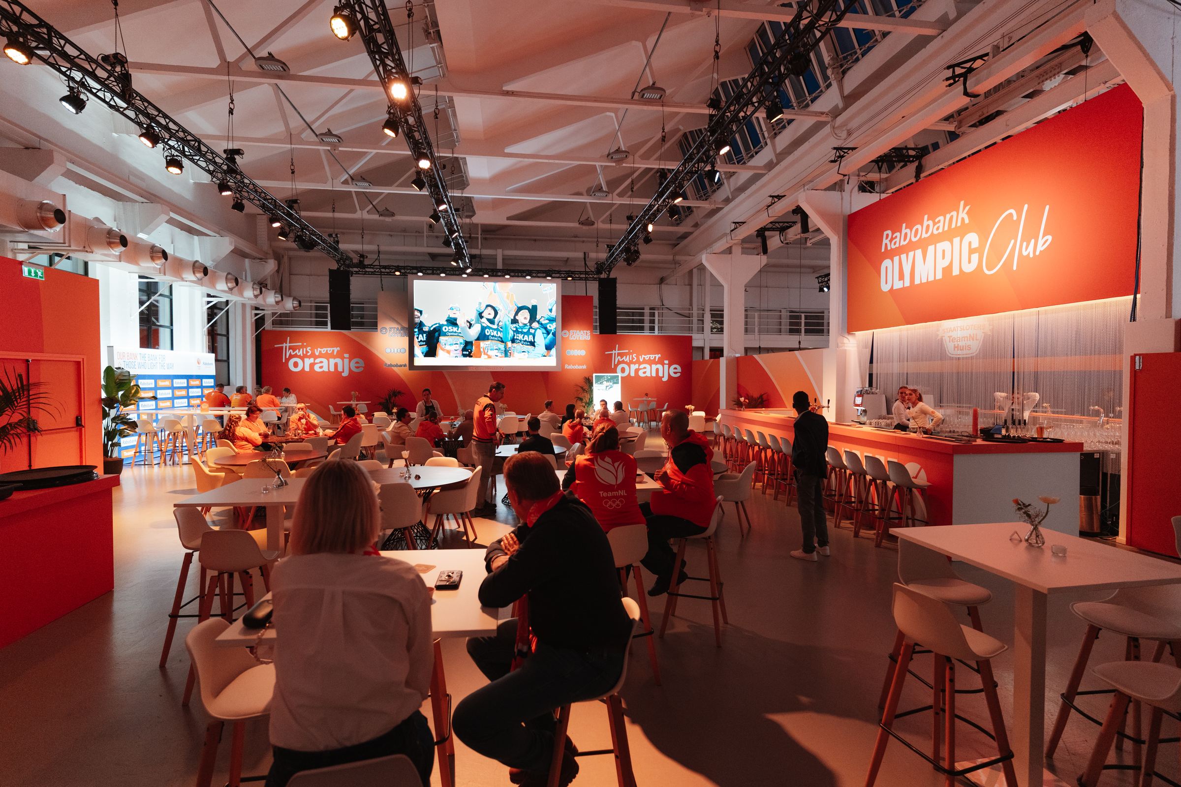 Zaal van het Staatsloterij TeamNL Huis in Milaan met bezoekers aan tafels, een groot scherm en de bar van de Rabobank Olympic Club